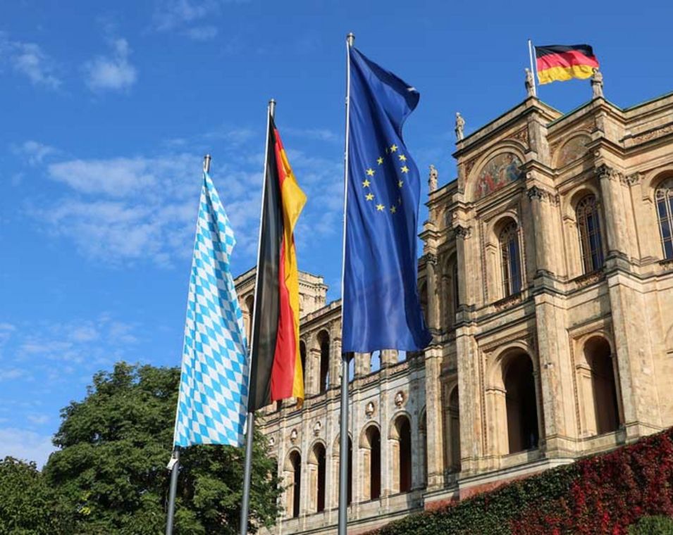 Ein historisches Gebäude mit der bayerischen, deutschen und europäischen Flagge davor. 
