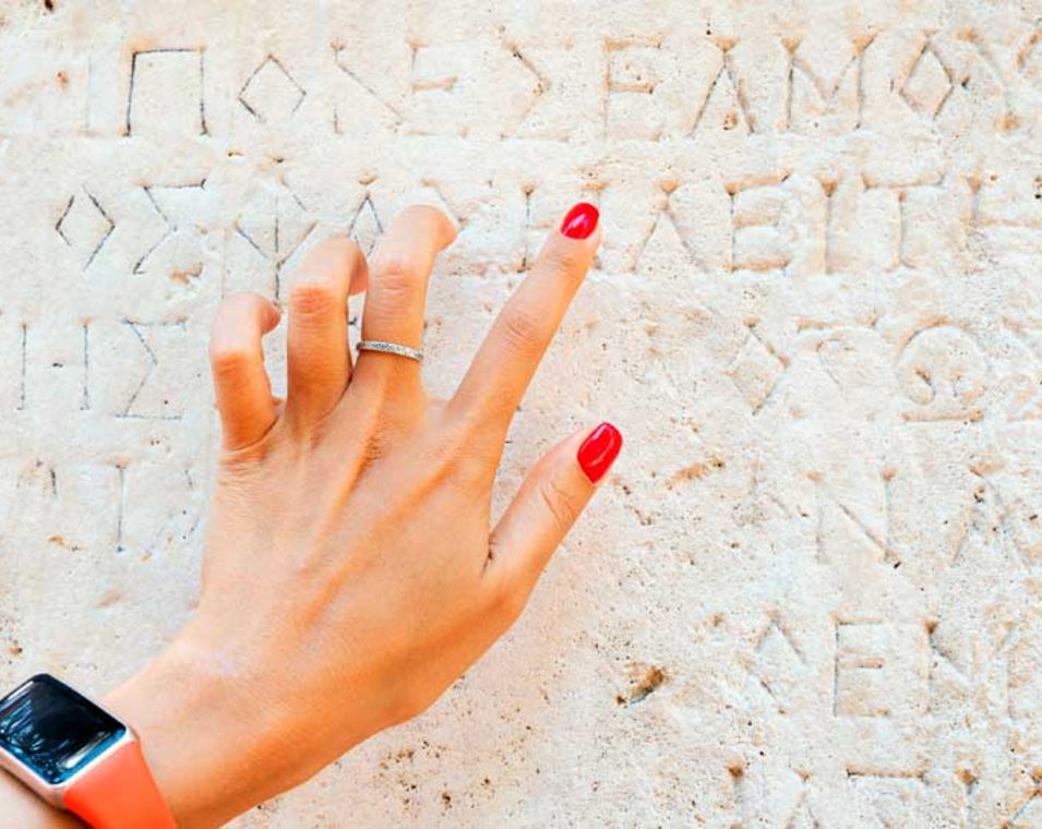 Hand einer jungen Frau, die auf die Schrift in einer antiken, griechischen Ruine zeigt und versucht die Schrift zu entziffern.