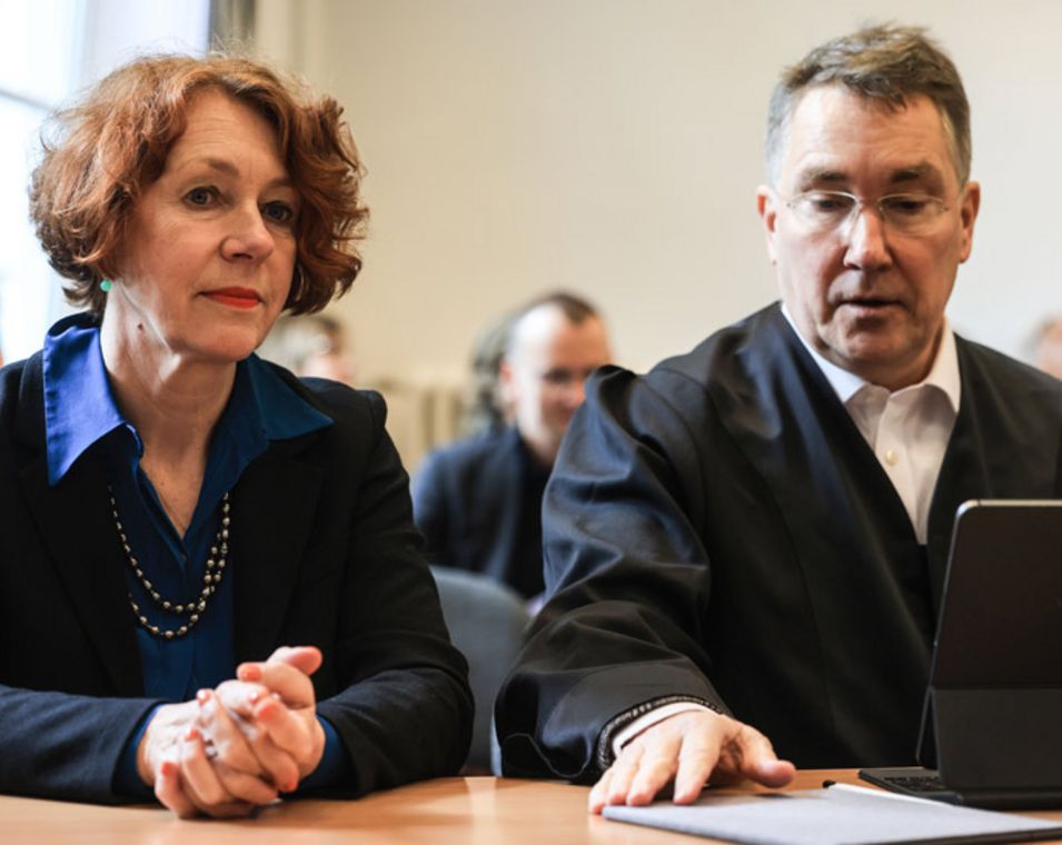 Eine Frau mit dunkelbraunem Haar und dunkelblauem Anzug sitzt in einem Gerichtssaal neben einem Mann in Anwaltsrobe. 