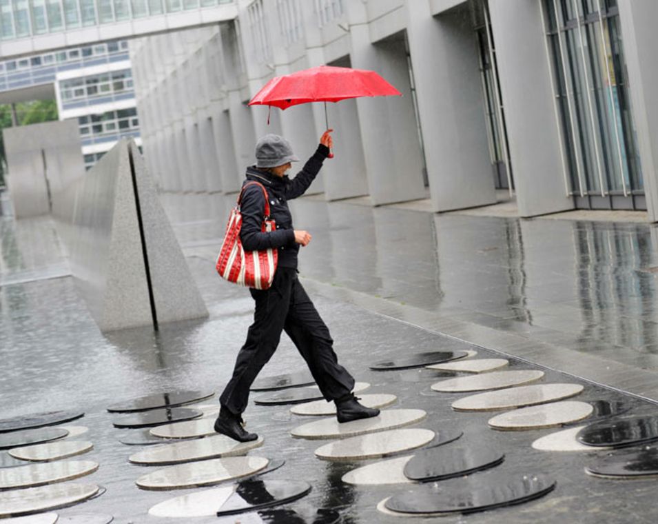 Eine Frau mit einem roten Schirm springt über die runden Metallplatten vor dem Europäischen Patentamt, ein großes, graues Bürogebäude. 