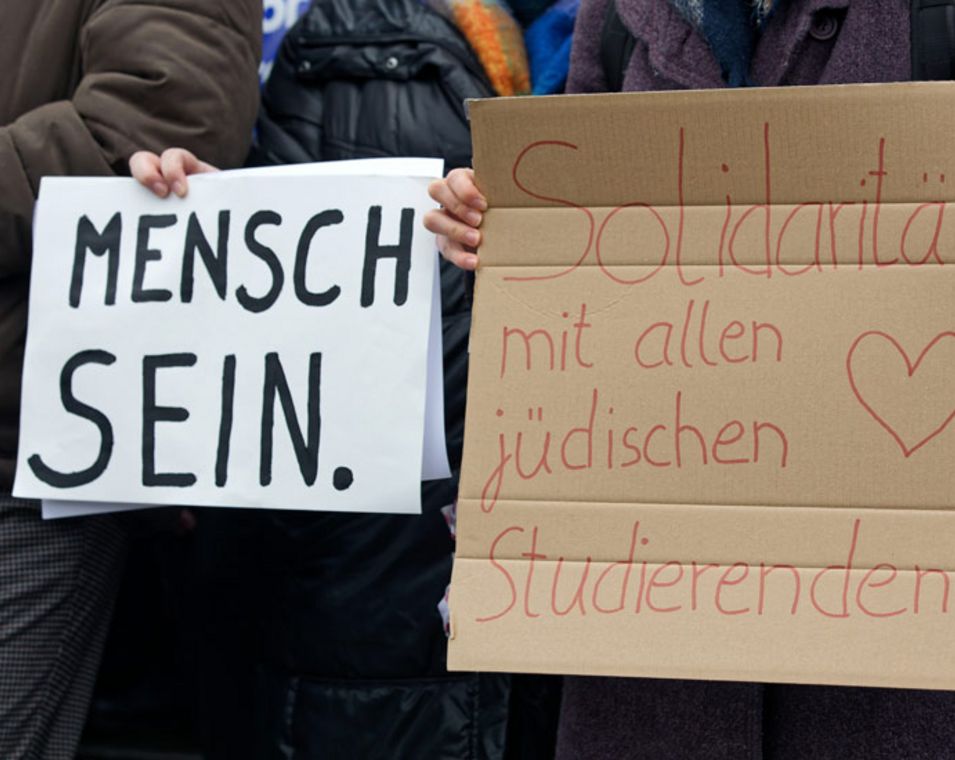 Auf zwei Protestbildern einer Demonstration ist zu lesen "MENSCH SEIN." und "Solidarität mit allen jüdischen Studierenden". 