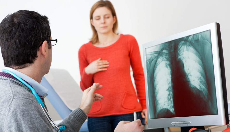 Ein Mann mit umgehängtem Stetoskop sitzt am Computer und betrachtet das Röntgenbild eine Patientin, die einige Schritte entfernt steht. 