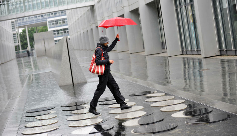 Eine Frau mit einem roten Schirm springt über die runden Metallplatten vor dem Europäischen Patentamt, ein großes, graues Bürogebäude. 