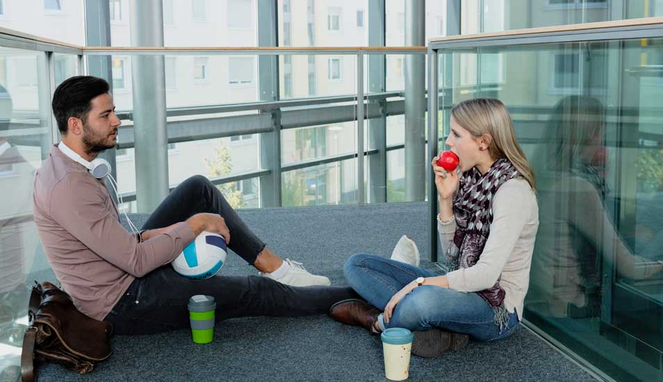 Junge Frau und junger Mann in modernem Bürogebäude machen Pause mit Kaffee, Apfel, Volleyball und Musik