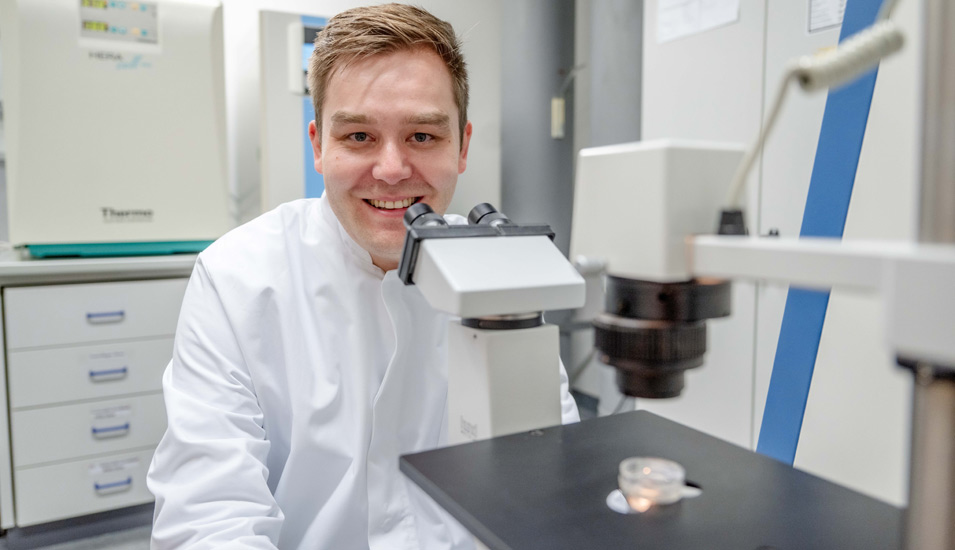 Ein junger Mann sitzt mit weißem Kittel in einem Labor hinter einem Mikroskop und lächelt.