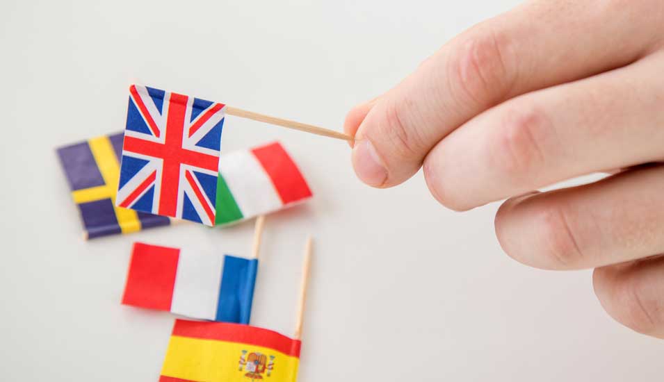 Eine Hand hält ein Fähnchen des Vereinigten Königreichs, auf dem Tisch darunter liegen weitere europäische Fähnchen, auch die spanische.
