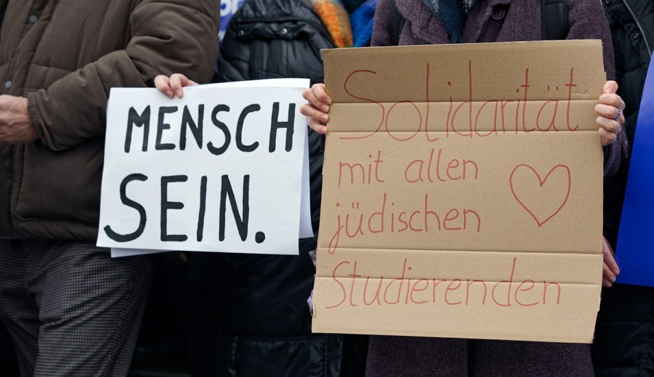 Auf zwei Protestbildern einer Demonstration ist zu lesen "MENSCH SEIN." und "Solidarität mit allen jüdischen Studierenden". 