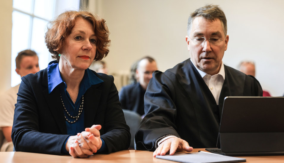 Eine Frau mit dunkelbraunem Haar und dunkelblauem Anzug sitzt in einem Gerichtssaal neben einem Mann in Anwaltsrobe. 