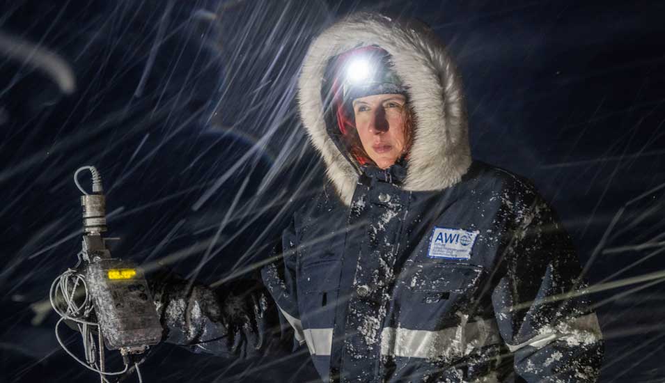 Wissenschaftlerin Julia Martin nimmt Messungen vor. Sie steht vor dunklem Hintergrund und trägt eine dicke Jacke mit Kapuze. Hauptsächlich ihr Gesicht ist beleuchtet, auf dem Kopf trägt sie eine Stirnlampe.