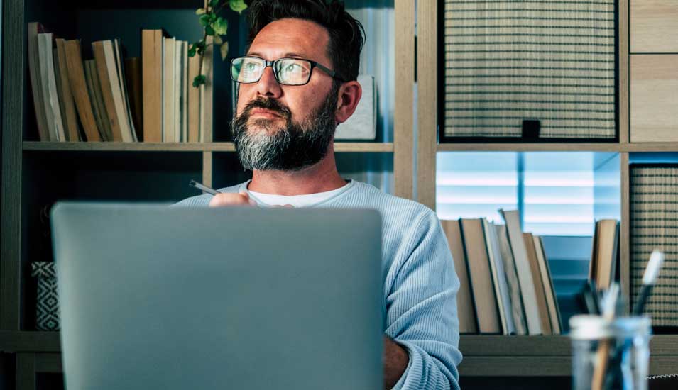 Mann mit Brille sitzt hinter einem Laptop am Schreibtisch und denkt nach, er hat die Augen vom Bildschirm abgewandt und blickt schräg nach oben.