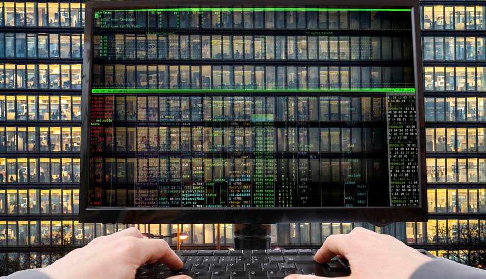 Durch einen Computerbildschirm hindurch ist ein Bürogebäude zu sehen und auf der Tastatur zwei Hände, die sich in das System einhacken. 
