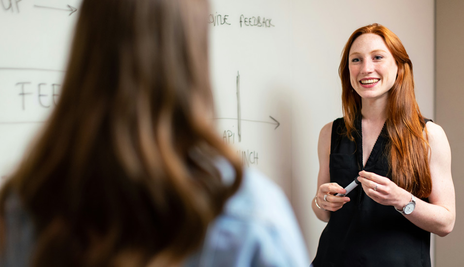 Eine Lehrerin in schwarzem Klein und mit rotblonden langen Haaren steht vor einer weißen Tafel im Gespräch mit einer jugendlichen Schülerin oder Studentin. 