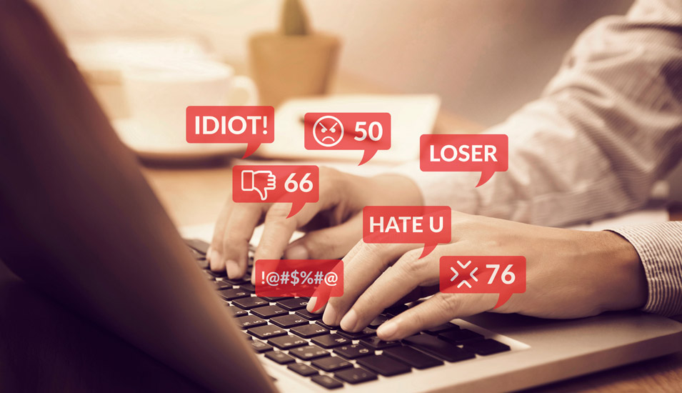 Hände tippen auf einer Laptop-Tastatur, aus welcher rote Lapel aufsteigen mit Hassrede-Begriffen und Emojis. 