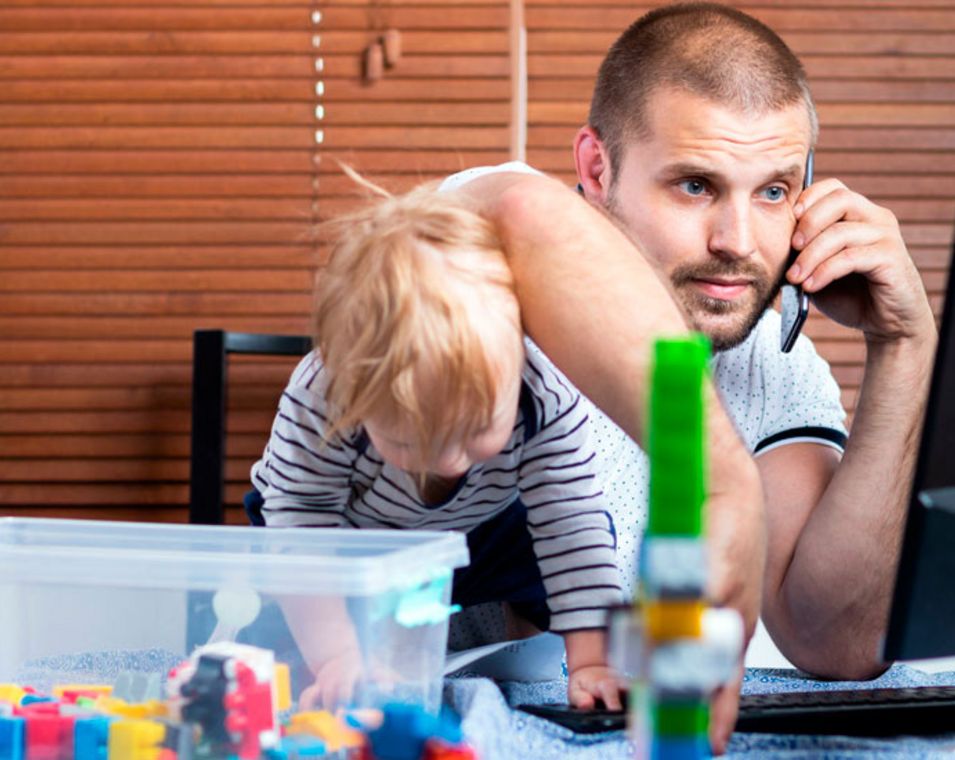 Ein Vater sitzt am Schreibtisch vor dem Computer und telefoniert, während sein Kind in seinen Armen turn und mit Legospielzeug auf dem Tisch spielt. 
