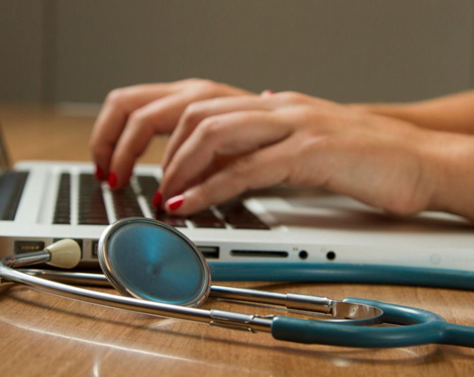 Frauenhände tippen auf einem Laptop an dessen Seite ein Stethoskop liegt. 
