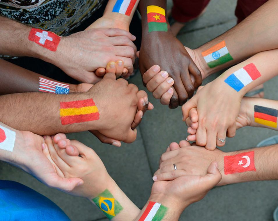Viele Hände mit aufgemalten Länder-Flaggen ergreifen einander freundlschaftlich in einem Kreis. 
