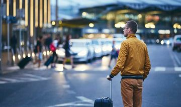 Junger Mann mit Koffer steht außerhalb eines Flughafen-Gebäudes