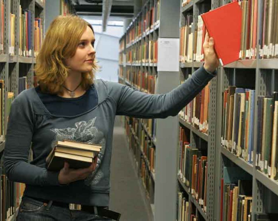 Das Foto zeigt eine junge Frau, die ein Buch aus einem Regal in einer Unibibliothek nimmt.