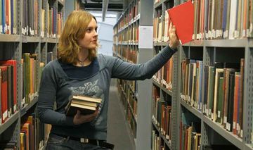 Das Foto zeigt eine junge Frau, die ein Buch aus einem Regal in einer Unibibliothek nimmt.