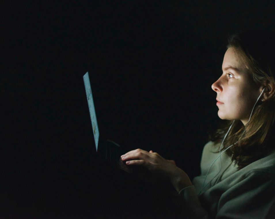 Eine junge Frau sitzt im Dunkeln vor ihrem Computer und tippt auf ihrer Tastatur. 