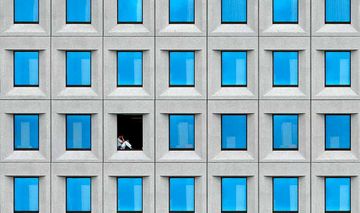 Gebäudefassade mit zahlreichen identischen geschlossenen Fenstern und einer Person, die in einem geöffneten Fenster sitzt