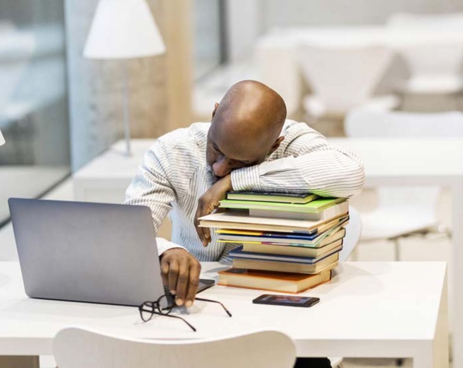 Mann mittleren Alters lehnt schlafend auf einem Stapel Bücher neben einem Laptop auf seinem Schreibtisch