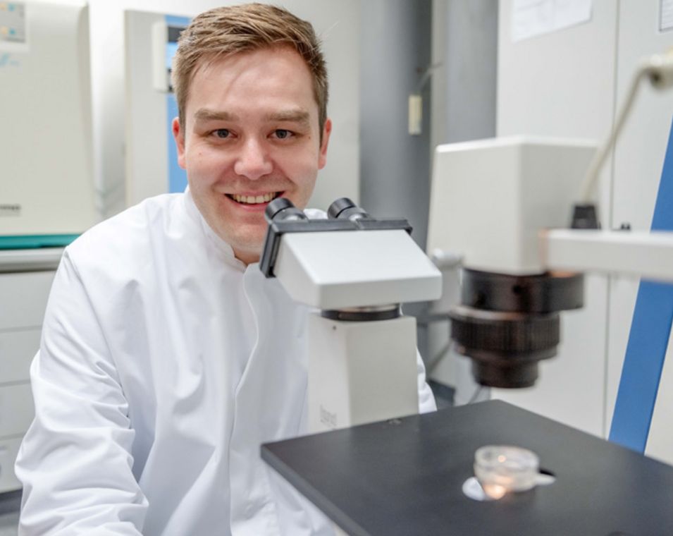 Ein junger Mann sitzt mit weißem Kittel in einem Labor hinter einem Mikroskop und lächelt.
