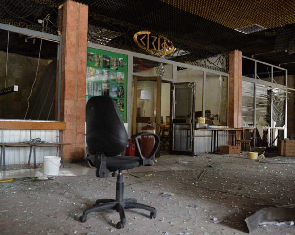 Das Bild zeigt die zerstörte Ausstattung der Hochschule in Charkiw mit Glassplittern auf dem Boden, zerstörtem Fenster und einem deplatzierten leeren Drehstuhl im Bildzentrum. 