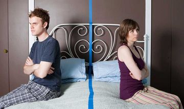 Ein Paar sitzt auf den Bettkanten eines Ehebettes und wendet sich jeweils die Rücken zu. Zwischen ihnen wurde mit Klebeband eine Trennlinie auf das Bett geklebt.