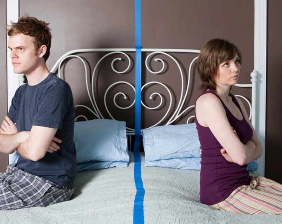 Ein Paar sitzt auf den Bettkanten eines Ehebettes und wendet sich jeweils die Rücken zu. Zwischen ihnen wurde mit Klebeband eine Trennlinie auf das Bett geklebt.
