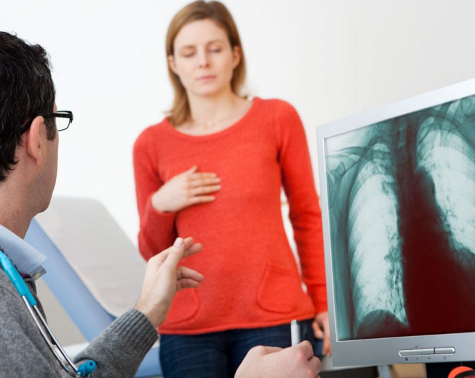 Ein Mann mit umgehängtem Stetoskop sitzt am Computer und betrachtet das Röntgenbild eine Patientin, die einige Schritte entfernt steht. 