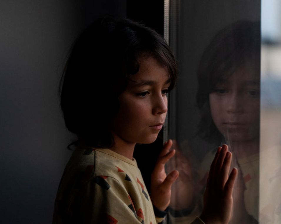 Ein Kind steht in einem dunklen Raum am Fenster und schaut nach draußen.