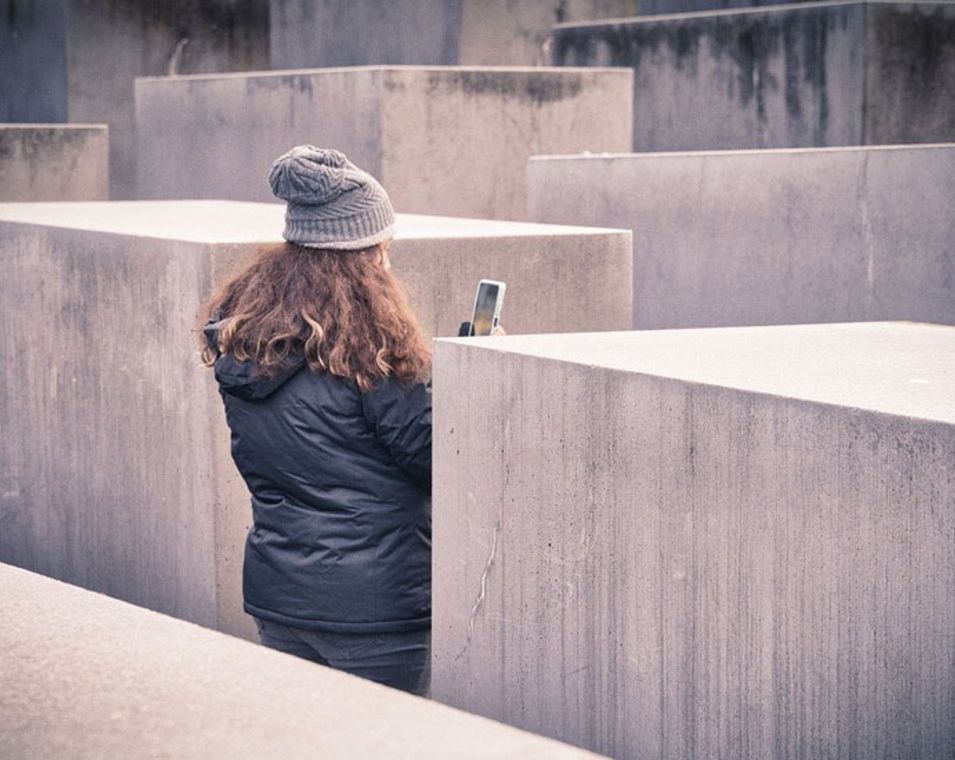 Eine junge Frau ist mit Mantel und Mütze von hinten zu sehen zwischen vielen grauen Steinquadern. Sie fotografiert mit ihrem Smartphone. 