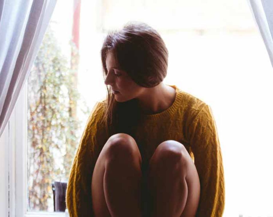 junge Frau sitzt mit angezogenen Beinen vor einem Fenster und schaut zur Seite