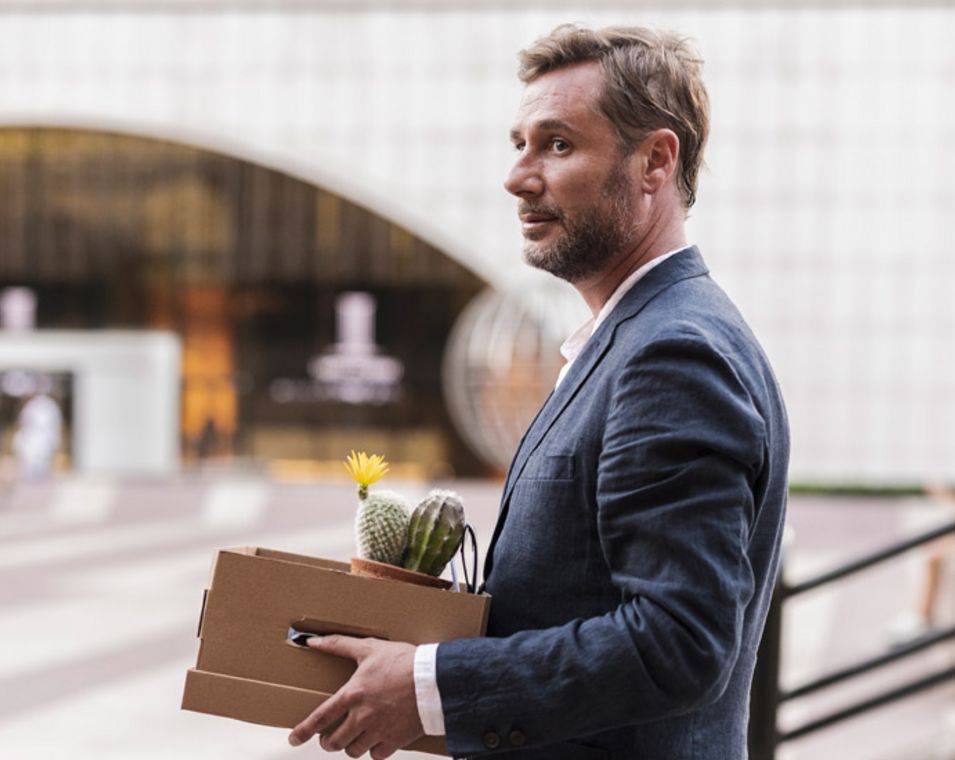 Ein Mann im Anzug vor einem großen Bürogebäude hät einen Karton in den Händen mit seinem persönlichen Utensilien vom Arbeitsplatz. 