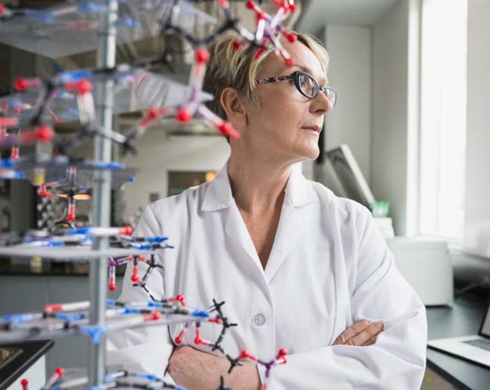 Forscherin steht vor einem DNA-Modell und schaut in Gedanken aus dem Fenster im Labor