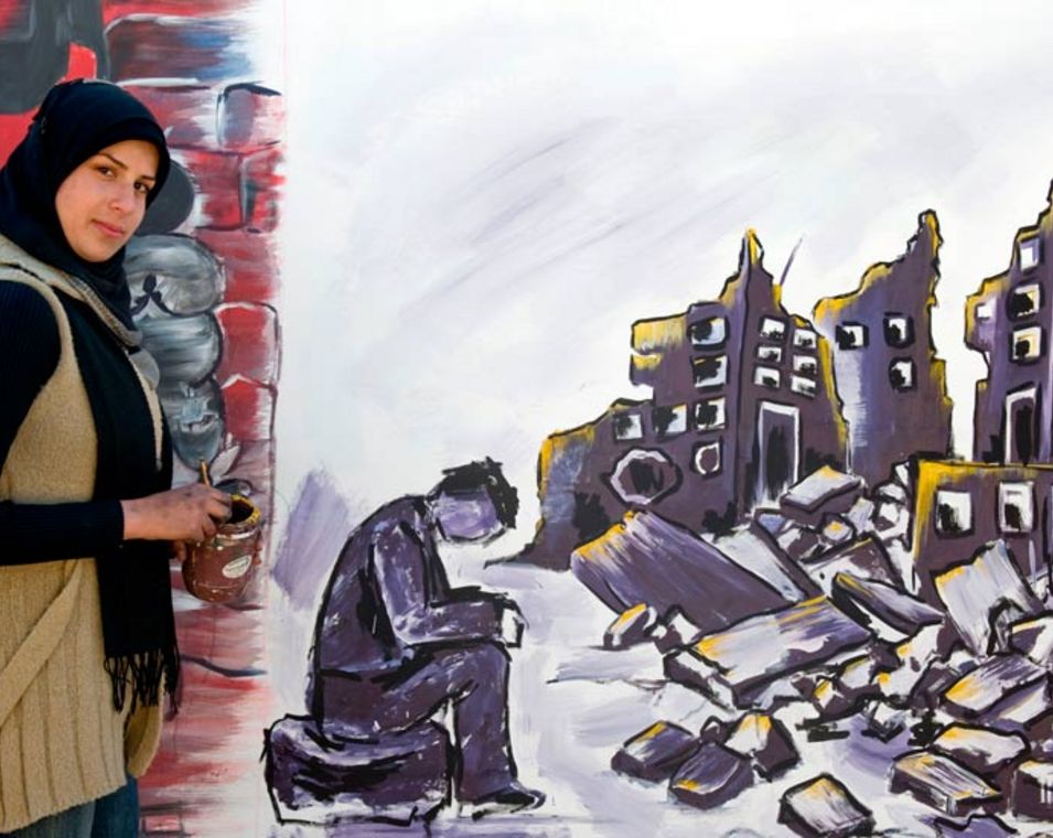 Eine junge Frau steht neben einer Häuserwand, auf die sie ein zerstörtes Gebäude gemacht hat in grau-schwarzen Farben. 