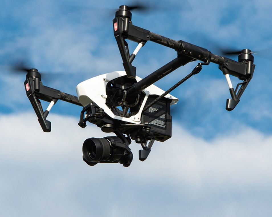 Eine Drohne mit integrierter Kamera fliegt vor einem leicht bewölkten Himmel. 