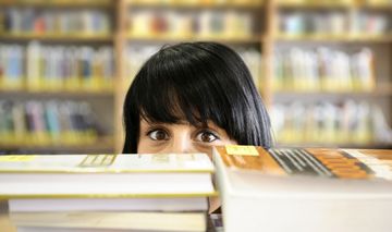 Das Foto zeigt eine Studentin hinter einem Bücherstapel, die in die Kamera schaut.