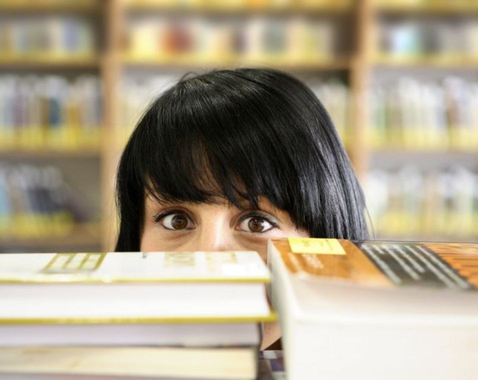 Das Foto zeigt eine Studentin hinter einem Bücherstapel, die in die Kamera schaut.