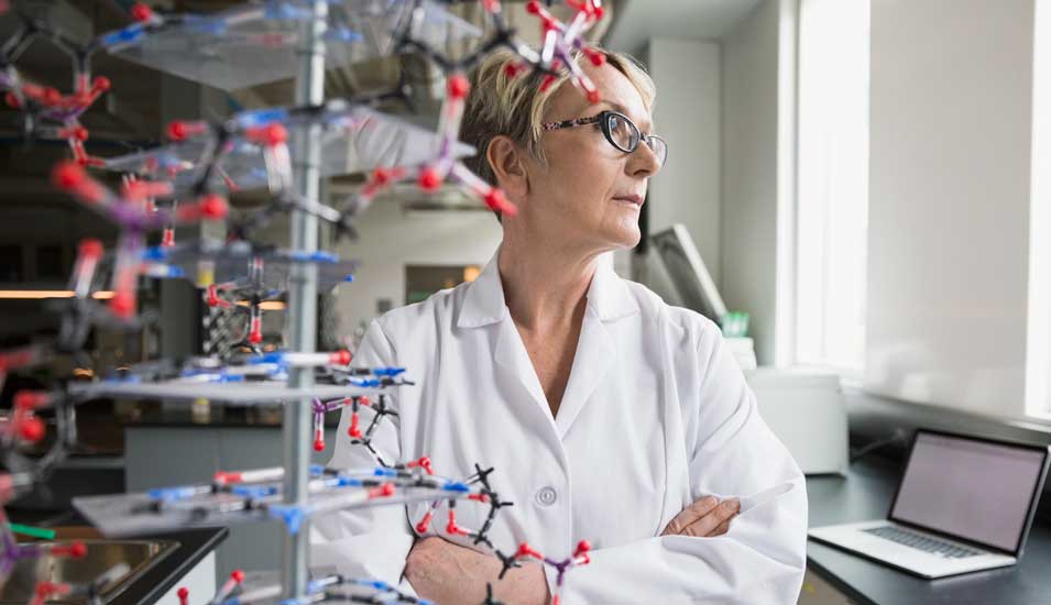 Forscherin steht vor einem DNA-Modell und schaut in Gedanken aus dem Fenster im Labor