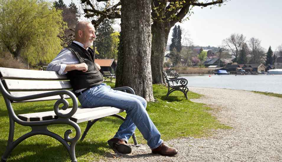 alter Mann mit grauem Bart sitzt auf einer Parkbank am See