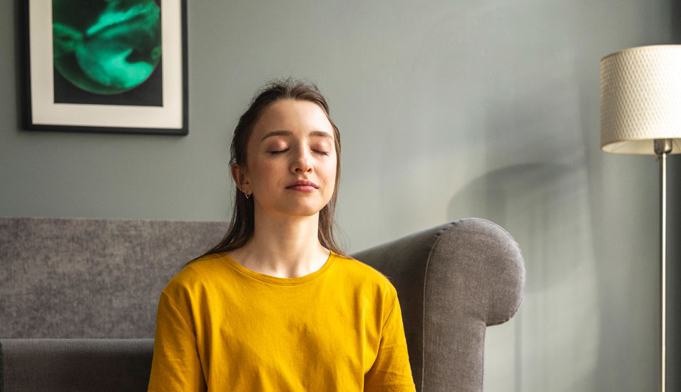 Eine junge Frau sitzt in ihrem Wohnzimmer und meditiert.