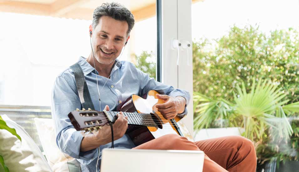 Mann übt Gitarre spielen mit Kopfhörern und einem Laptop in einem Wohnzimmer