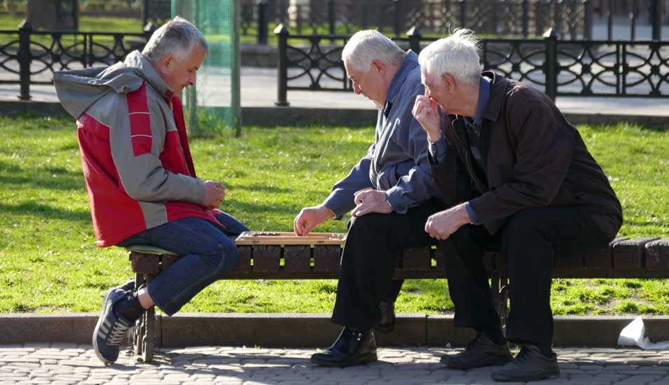 Drei ältere Männer sitzen auf einer Parkbank und spielen Backgammmonn
