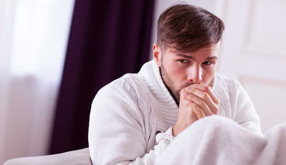 Junger Mann sitzt in dickem Pullover und mit einer Decke auf einem Sofa und friert