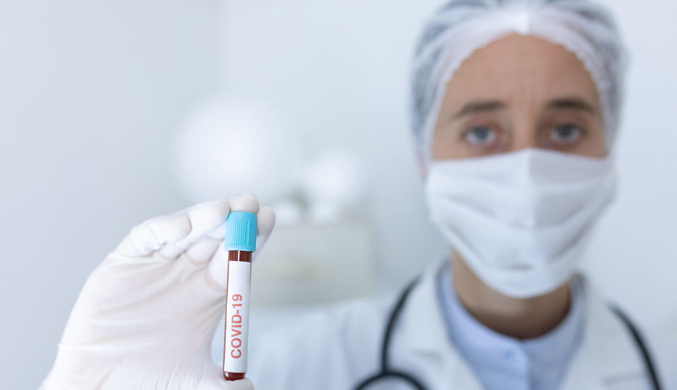 Eine Ärztin mit Maske und Op-Haube hält eine Blutprobe in die Kamera, auf der Blutprobe steht "Covid-19".