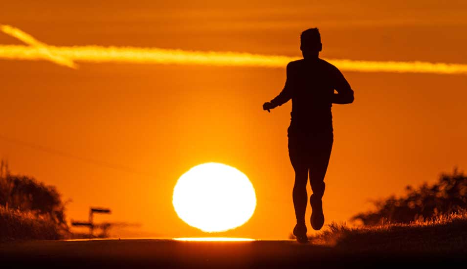 Ein Mann läuft bei Sonnenaufgang einen Feldweg entlang. Der Himmel ist orange gefärbt.