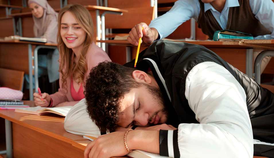Student schläft mit Kopf auf den Armen in einem Hörsaal, Mitstudenten beobachten ihn