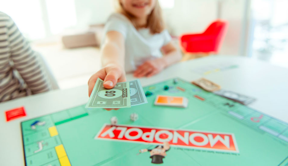 Kind spielt das Brettspiel "Monopoly" und reicht Geldscheine in Richtung des Bildbetrachters.